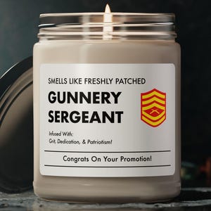 May include: A lit candle in a glass jar with a silver lid. The label reads "Smells Like Freshly Patched Gunner Sergeant" with a military insignia and the text "Congrats On Your Promotion!" The candle is on a dark surface.