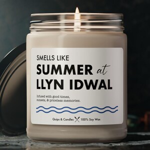 May include: A glass jar candle with a silver lid, lit with a flame. The label says "SMELLS LIKE SUMMER at LLYN IDWAL" with a blue wavy line. The candle is infused with good times, sunsets, and priceless memories.