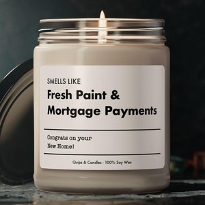 May include: A beige candle with a black label that reads "SMELLS LIKE Fresh Paint & Mortgage Payments Congrats on your New Home! Quips & Condles 100% Soy Wax".
