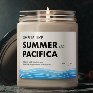 May include: A lit candle in a glass jar with a silver lid. The label reads "SMELLS LIKE SUMMER in PACIFICA" with a blue wave design and the text "Infused with good times, sunshine, & priceless memories."