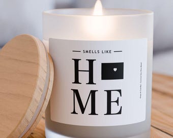 Op de afbeelding: Een frosted glazen kaars met een wit label waarop "Smells Like Home" staat met een zwart hart in de "O". De kaars heeft een houten deksel en staat op een houten tafel.