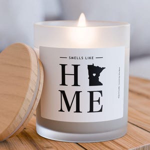 May include: A white frosted glass candle with a black and white label that says "Smells Like Home" with an outline of the state of Minnesota. The candle is lit and has a wooden lid.