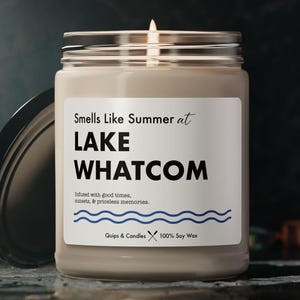 Könnte beinhalten: Ein braunes Glasgefäß mit einer Kerze und einem weißen Etikett mit der Aufschrift "Smells Like Summer at LAKE WHATCOM" und "Infused with good times, tunes, & precious memories." Das Etikett enthält auch den Text "Quips & Candles 100% Soy Wax".