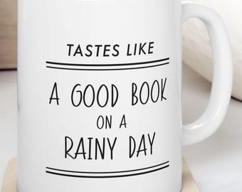 May include: White ceramic mug with the text "TASTES LIKE A GOOD BOOK ON A RAINY DAY" in black. The mug is on a beige coaster. The background includes a wooden cutting board, a bowl with fruit, and a white surface.