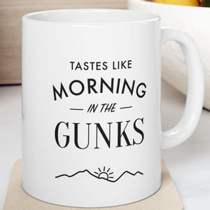 May include: White ceramic coffee mug with black text that reads "Tastes like morning in the gunks" with a small mountain and sun graphic below the text.