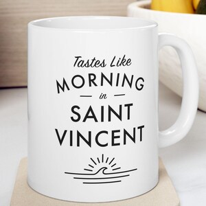 May include: White ceramic mug with black text that reads "Tastes Like Morning in Saint Vincent" and a black silhouette of a sun and wave.