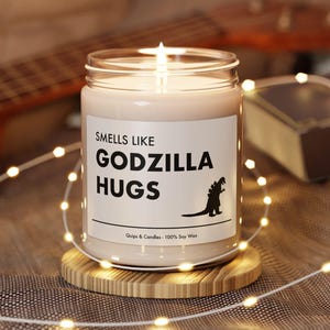 May include: A white candle in a clear glass jar with a black label that reads "Smells Like Godzilla Hugs". The candle is on a wooden coaster and surrounded by white fairy lights.