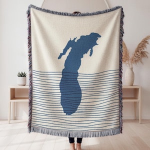 May include: A woven throw blanket featuring a cream-colored base with a large, solid dark blue silhouette of a lake shape centered in the middle. The lower half of the blanket displays horizontal dark blue wavy lines, and the edges have fringe.