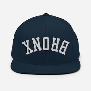 Puede incluir: Gorra de béisbol azul marino con la palabra "BRONX" bordada en blanco, arqueada en la parte delantera. La gorra tiene una visera plana y una corona estructurada. Un sombrero de estilo clásico, adecuado para ropa informal.
