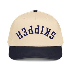 May include: A baseball cap with a cream-colored crown and a navy blue brim. The word "SKIPPER" is embroidered in navy blue arched lettering across the front of the cap. The cap has a black button on top.