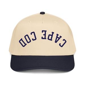 Puede incluir: Gorra de béisbol con corona color crema y visera azul marino. El texto "CAPE COD" está bordado en azul marino en la parte delantera. La gorra tiene un botón azul marino oscuro en la parte superior.