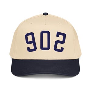 Pode incluir: Boné de beisebol com coroa creme e aba azul marinho. O número "902" é bordado em azul marinho na frente. O boné tem um botão preto na parte superior.