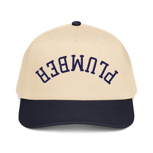 May include: A baseball cap with a cream-colored crown and a navy blue brim. The word "PLUMBER" is embroidered in navy blue across the front of the cap. The cap has a black button on top.