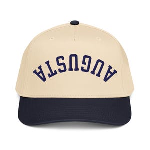 Può includere: Un berretto da baseball con corona color crema e visiera blu navy. La parola "AUGUSTA" è ricamata in lettere arcuate blu navy sul davanti. Il berretto ha un bottone nero sulla parte superiore.