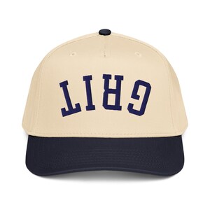 May include: A baseball cap with a cream-colored crown and a navy blue brim. The word "GRIT" is embroidered in navy blue block letters across the front of the cap. The cap has a black button on top.
