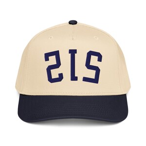 Pode incluir: Boné de beisebol com coroa creme e aba azul marinho. O boné apresenta o número "215" em letras de bloco azul marinho na frente. O boné tem uma aba curva e um botão na parte superior.