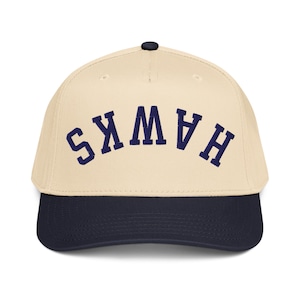 May include: A baseball cap with a cream-colored crown and a navy blue brim. The word "HAWKS" is arched across the front in navy blue block letters. The cap has a black button on top.
