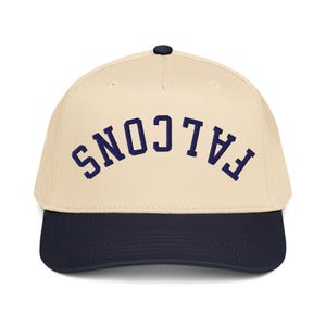 Può includere: Cappellino da baseball beige con visiera blu navy e bottone blu navy. La parola "FALCONS" è arcuata sulla parte anteriore in lettere blu navy.