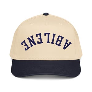 Op de afbeelding: Een baseballpet met een crèmekleurige kroon en een marineblauwe klep. Het woord "ABILENE" staat in marineblauwe letters in een boog over de voorkant. De pet heeft een zwarte knop bovenop.