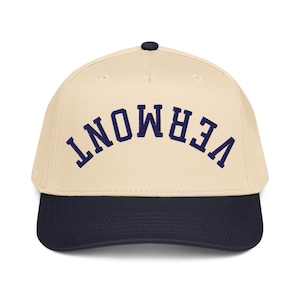 Puede incluir: Gorra de béisbol con corona color crema y visera azul marino. La palabra "VERMONT" está bordada en azul marino en la parte delantera, con letras arqueadas. La gorra tiene un botón negro en la parte superior.