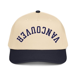 May include: A baseball cap with a cream-colored crown and a navy blue brim. The word "VANCOUVER" is arched across the front in navy blue lettering. The cap has a dark navy button on top.