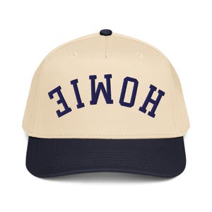 May include: A baseball cap with a cream-colored crown and a navy blue brim. The word "HOMIE" is embroidered in navy blue block letters across the front of the cap. The cap has a curved brim and a black button on top.