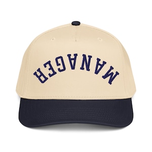 May include: A baseball cap with a cream-coloured crown and a navy blue brim. The word "MANAGER" is arched across the front in navy blue lettering. The cap has a black button on top.