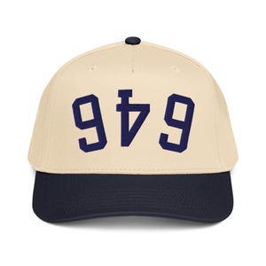 Pode incluir: Boné de beisebol com coroa creme e aba azul marinho. O boné apresenta o número "946" em letras de bloco azul marinho na frente. O boné tem um botão preto na parte superior.