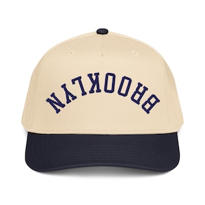 May include: A baseball cap with a cream-colored crown and a navy blue brim. The word "BROOKLYN" is embroidered across the front in navy blue, arched lettering. The cap has a black button on top.