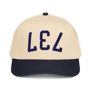 Pode incluir: Boné de beisebol com coroa creme e aba azul marinho. O boné apresenta as letras "LEZ" em azul marinho em uma fonte em negrito sem serifa.