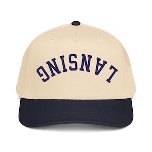 Peut inclure: Casquette de baseball avec une couronne crème et une visière bleu marine. Le mot "LANSING" est brodé en bleu marine en arc sur le devant de la casquette. La casquette a un bouton noir sur le dessus.