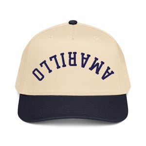 May include: A baseball cap with a cream-colored crown and a navy blue brim. The word "AMARILLO" is embroidered in navy blue across the front of the cap.