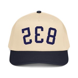 Pode incluir: Boné de beisebol com coroa creme e aba azul marinho. O boné apresenta o número "238" em letras de bloco azul marinho na frente. O boné tem um botão preto na parte superior.