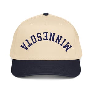 May include: A baseball cap with a cream-colored crown and a navy blue brim. The word "MINNESOTA" is embroidered in navy blue across the front of the cap in a curved, arched design. The cap has a black button on top.