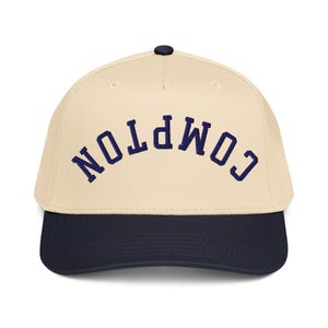 May include: A baseball cap with a cream-colored crown and a navy blue brim. The word "COMPTON" is embroidered across the front in navy blue, arched lettering. The cap has a black button on top.