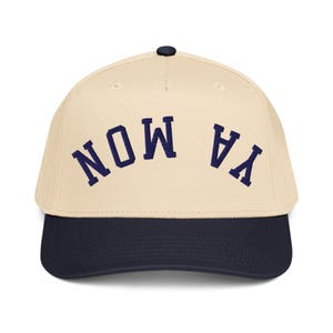 May include: A baseball cap with a cream-colored crown and a navy blue brim. The cap features the words "NOW YA" in navy blue arched across the front. The cap has a black button on top.