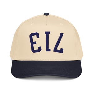 May include: A baseball cap with a cream-coloured crown and a navy blue brim. The cap features the number "713" in navy blue block letters on the front. The cap has a black button on top.