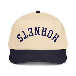 May include: A cream-coloured baseball cap with a navy blue brim and the navy blue text "HORNETS" arched across the front. The cap has a curved brim and a small black button on top. This hat is a classic style.