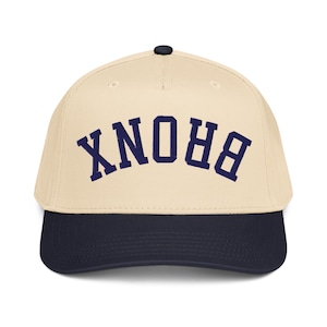 Puede incluir: Gorra de béisbol con corona color crema y visera azul marino. La palabra "BRONX" está bordada en letras mayúsculas azul marino en la parte delantera. La gorra tiene una visera curva y un pequeño botón negro en la parte superior.