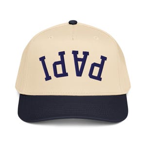 May include: A baseball cap with a cream-colored crown and a navy blue brim. The word "PAPI" is embroidered in navy blue across the front of the cap.