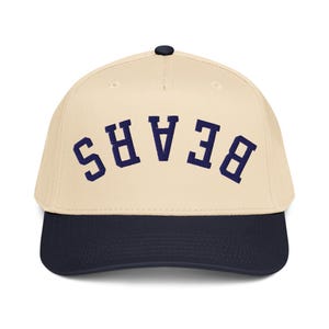 Könnte beinhalten: Baseballkappe mit cremefarbener Krone und marineblauem Schirm. Das Wort "BEARS" ist in Marineblau auf die Vorderseite gestickt. Die Kappe hat einen gebogenen Schirm und einen kleinen schwarzen Knopf oben.