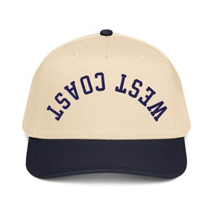 May include: A baseball cap with a cream-colored crown and a navy blue brim. The arched text "WEST COAST" is embroidered in navy blue across the front. The cap has a dark navy button on top.