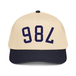 Pode incluir: Boné de beisebol com coroa creme e aba azul marinho. O boné apresenta o número "984" em letras de bloco azul marinho na frente. O boné tem uma aba curva e um botão na parte superior.