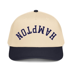 May include: A baseball cap with a cream-colored crown and a navy blue brim. The word "HAMPTON" is embroidered in navy blue arched across the front. The cap has a black button on top.