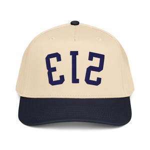 Pode incluir: Boné de beisebol com coroa creme e aba azul marinho. O boné apresenta o número "213" em letras de bloco azul marinho na frente. Possui um botão preto na parte superior.