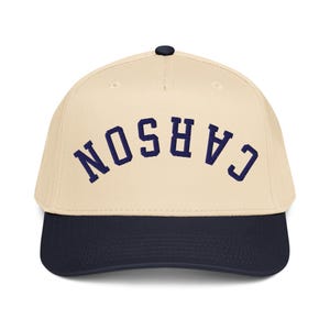 Puede incluir: Gorra de béisbol con corona color crema y visera azul marino. La palabra "CARSON" está bordada en azul marino en la parte delantera. La gorra tiene un botón negro en la parte superior.