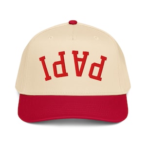 May include: A cream-coloured baseball cap with a red brim and a red button on top. The front of the cap features the word PAPI embroidered in red block letters, positioned in an upside-down, arched orientation across the centre of the crown.