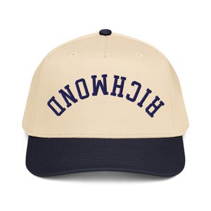 May include: A baseball cap with a cream-colored crown and a navy blue brim. The word "RICHMOND" is embroidered in navy blue arched lettering across the front of the cap. The cap has a black button on top.