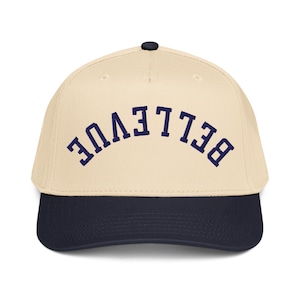 May include: A baseball cap with a cream-colored crown and a navy blue brim. The word "Bellevue" is embroidered in navy blue across the front of the cap. The cap has a black button on top.