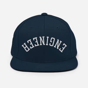May include: A navy blue flat-brimmed baseball cap featuring the word "ENGINEER" embroidered in white, curved across the front. The cap has a structured fit and a rounded crown. A classic accessory for those in the engineering field.